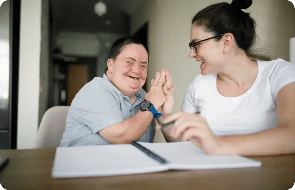 A Jango Community Services support worker and a young man with Down syndrome laughing and high-fiving over an open notebook, showcasing a meaningful connection and goal-oriented support.
