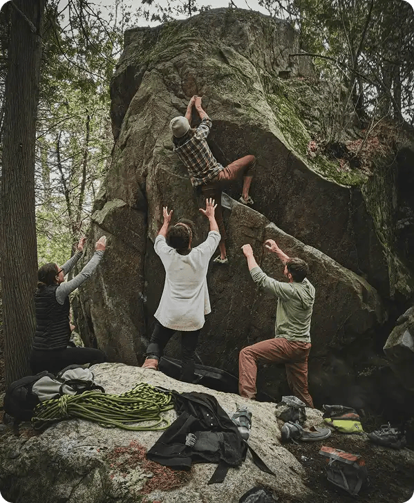 A person in a plaid shirt climbs a large rock while three others stand below with their hands raised to "spot" and support them, symbolizing Jango’s role in coordinating a network of providers to ensure the safety and success of NDIS participants.