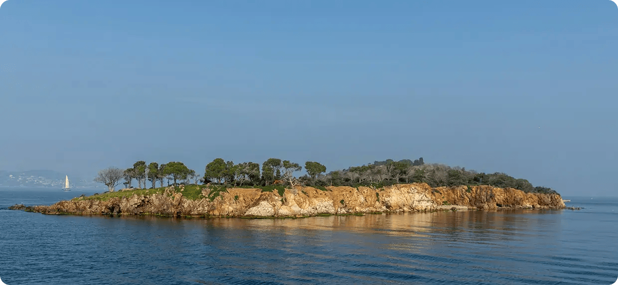 A wide scenic view of a rocky, tree-lined island in calm blue waters with a small sailboat in the distance, representing the peaceful and restorative coastal environment where Jango provides local NDIS support services in Hervey Bay.