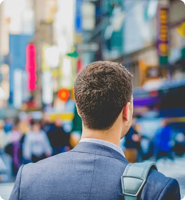 A view from behind a young man in a professional grey suit and backpack, looking toward a bustling city street, representing the final stage of Jango's Pathways to Potential program where participants confidently enter the workforce and broader community.