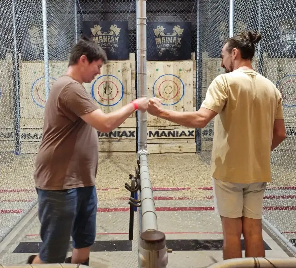 Two men fist bump inside an indoor axe-throwing venue at Jango’s, standing in adjacent lanes with wooden targets mounted against a fenced backdrop.