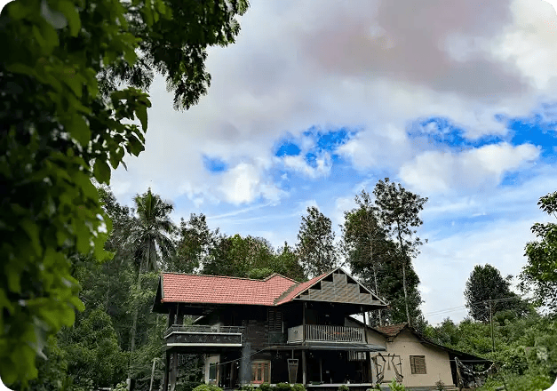 A large, modern two-story home with a red tile roof surrounded by lush green trees under a bright cloudy sky, representing Jango’s high-quality residential Short Term Accommodation options that provide a safe and welcoming "home away from home" for NDIS participants.
