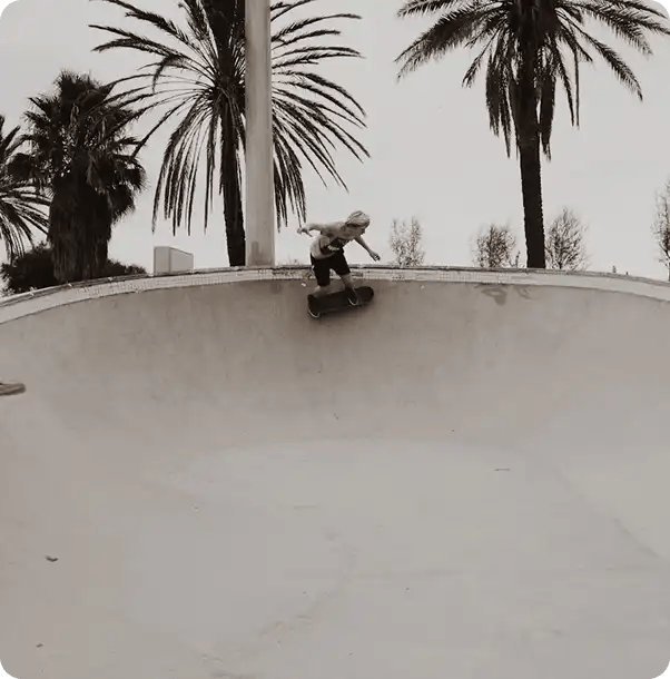 A black and white photo of a person skateboarding along the rim of a concrete skate bowl under palm trees, representing Jango’s support for youth NDIS participants in pursuing active hobbies and social inclusion.