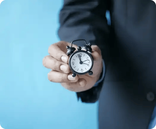 A close-up of a person in a professional suit holding a tiny black alarm clock against a blue background, symbolizing Jango’s commitment to time management, efficiency, and respecting the work-life balance of our team members.