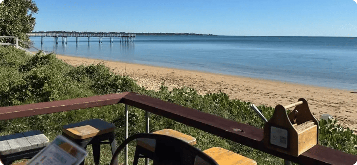 A peaceful view from a wooden deck overlooking a golden sand beach and a long pier stretching into calm blue water, representing the relaxing and high-quality respite environments provided by Jango’s STA services.