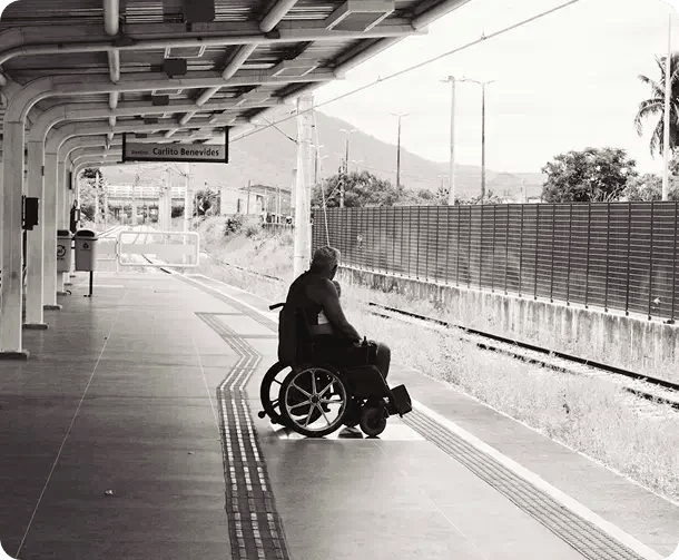 A black and white photograph of a person in a wheelchair waiting on a train station platform, symbolizing Jango’s commitment to providing accessible disability support and transport assistance throughout Queensland and Northern NSW.